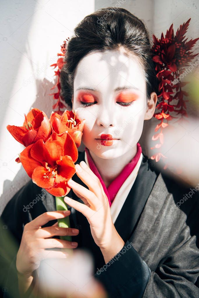 selective focus of sakura branches and beautiful geisha holding red flowers with closed eyes in sunlight