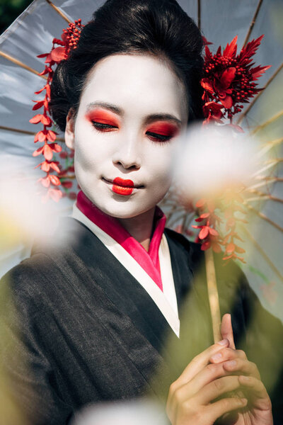 selective focus of tree branches and beautiful smiling geisha with umbrella and closed eyes in sunlight