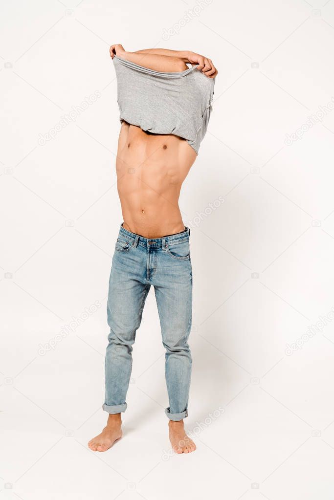 muscular and shirtless man covering face while taking off grey t-shirt on white 