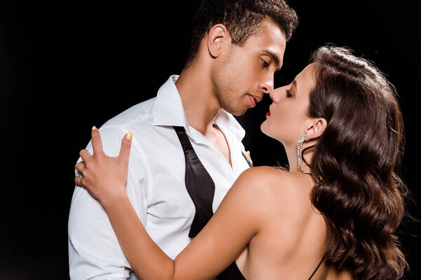 handsome mixed race man hugging and looking at elegant woman isolated on black 