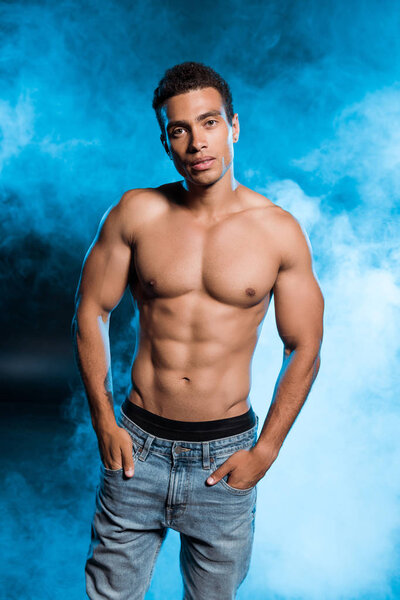 sexy mixed race man standing with hands in pockets and looking at camera on blue with smoke 