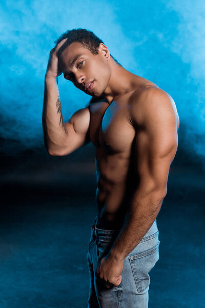 mixed race man with tattoo touching hair on blue with smoke 
