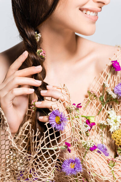 cropped view of smiling woman touching braid in mesh with spring wildflowers isolated on grey