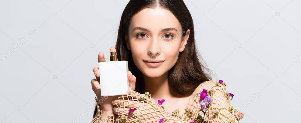beautiful smiling woman in mesh clothing with purple flowers holding perfume bottle with copy space and floral scent isolated on grey