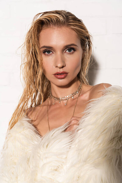 sexy naked young woman with wet hair in white faux fur coat looking at camera on white background