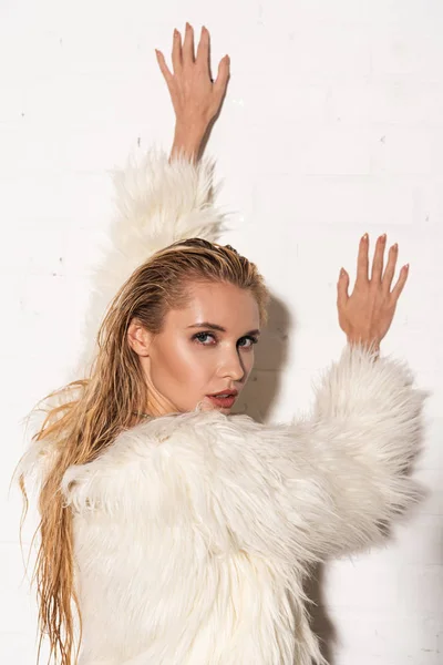 sexy young woman with wet hair in white faux fur coat posing near white brick wall