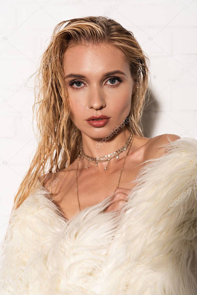 sexy naked young woman with wet hair in white faux fur coat looking at camera on white background
