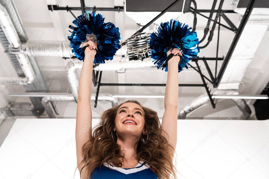 bottom view of happy smiling cheerleader girl in blue uniform dancing with pompoms