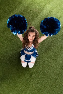 top view of sexy smiling cheerleader girl in blue uniform sitting with pompoms on green field clipart