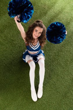 top view of sexy cheerleader girl in blue uniform sitting with pompoms on green field clipart