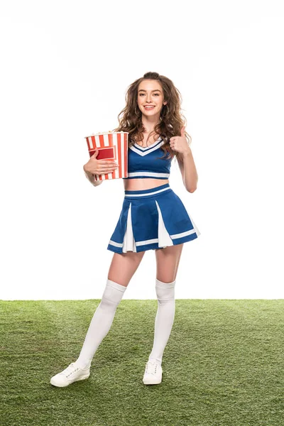快乐啦啦队长女孩在蓝色制服站在一桶爆米花 并显示大拇指在绿色领域隔离在白色 — 图库照片
