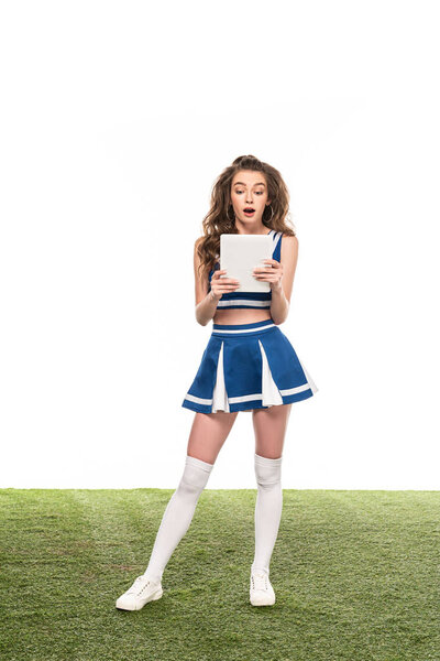 shocked cheerleader girl in blue uniform holding digital tablet on green grass isolated on white