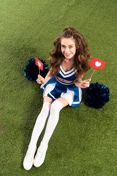 overhead view of pretty cheerleader girl in blue uniform sitting with pompoms and social media hearts on green field