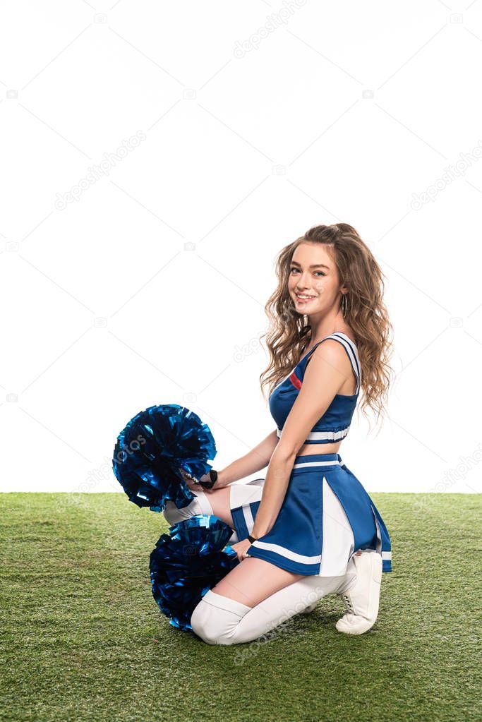 side view of sexy happy cheerleader girl in blue uniform with pompoms sitting on grassy field isolated on white