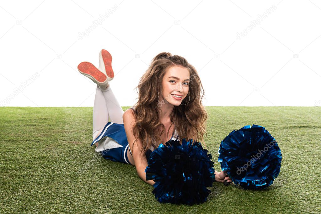 smiling cheerleader girl in blue uniform lying with pompoms on green field isolated on white