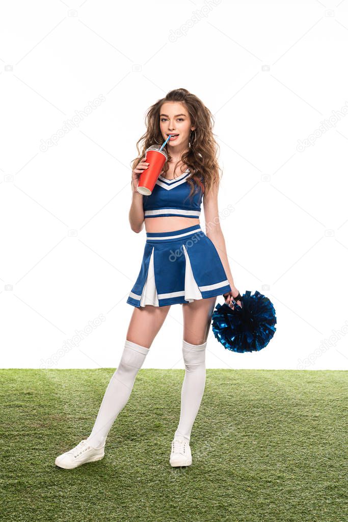 sexy cheerleader girl in blue uniform standing with soda and pompom on green field isolated on white