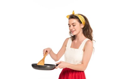 elbise ve önlük tutan tava ve ahşap spatula beyaz izole genç gülümseyen ev kadını