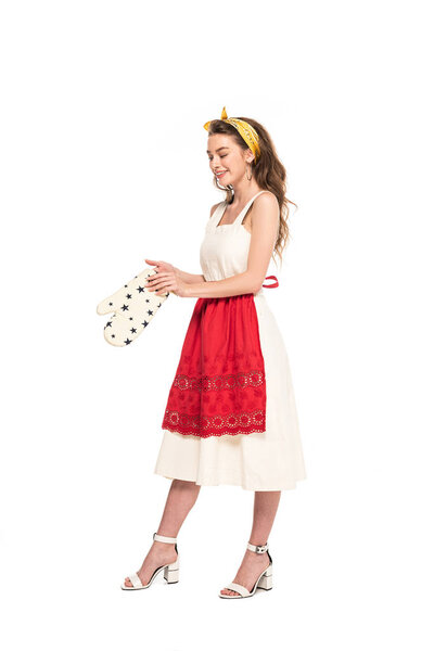 full length view of young happy housewife in dress and apron holding oven mitten isolated on white