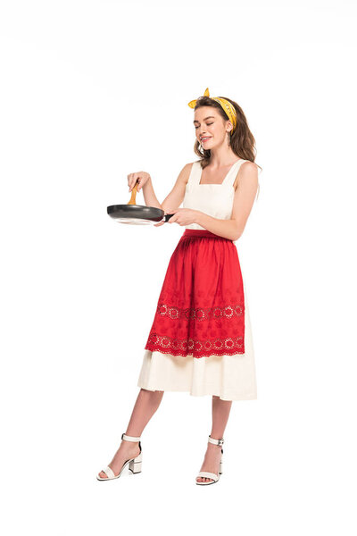 full length view of young housewife in dress and apron holding wooden spatula and frying pan isolated on white