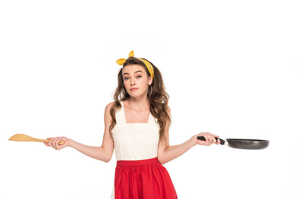 young housewife in dress and apron holding frying pan and wooden spatula while showing shrug gesture isolated on white