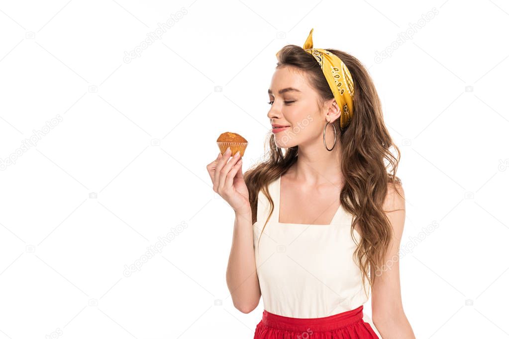 young pleased housewife in dress and apron looking at muffin isolated on white
