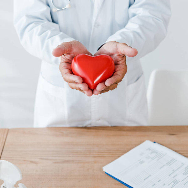 cropped view of doctor holding red heart model near table 