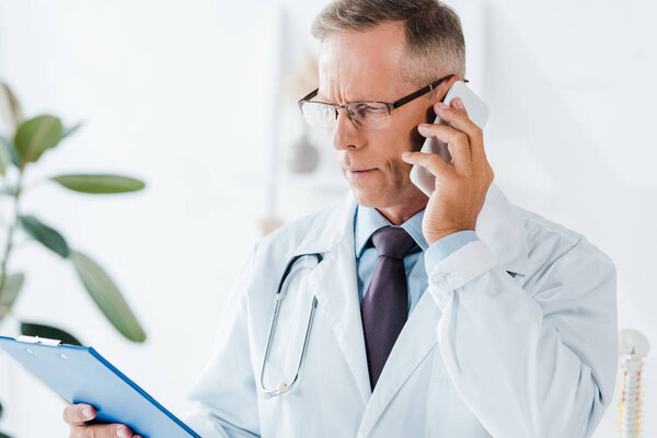 serious doctor in glasses looking at clipboard while talking on smartphone 