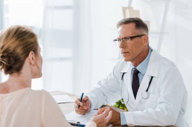 beyaz ceket Doktor seçici odak kadın bakıyor ve klinikte kalem tutarak 