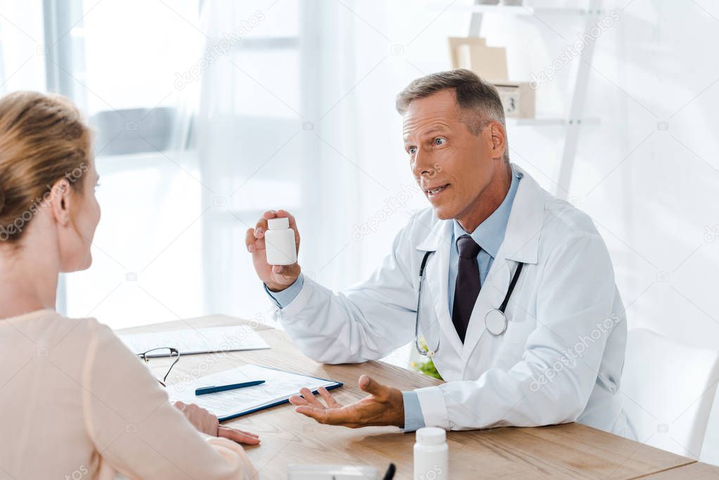doctor in white coat sitting and gesturing while holding bottle and looking at woman 