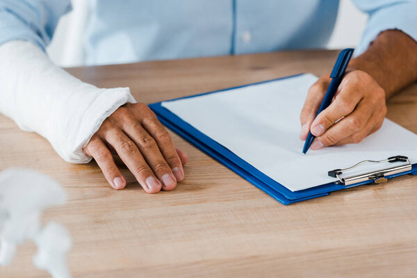 cropped view of injured man with gypsum bandage writing on clipboard in hospital 