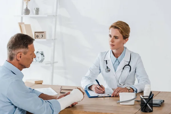 Attractive Doctor Looking Patient Broken Arm While Writing Diagnosis Clinic — Stock Photo, Image