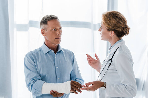 привлекательный врач жестом рядом с мужчиной со сломанной рукой в клинике
 