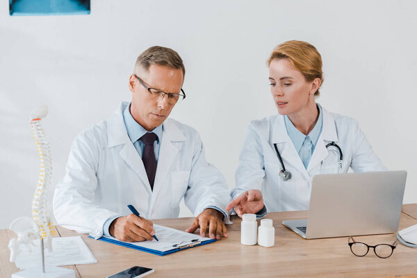 attractive doctor pointing with finger near laptop and coworker in glasses writing diagnosis 