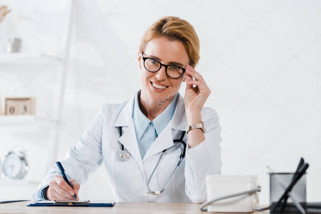happy doctor touching glasses and writing diagnosis in clinic 