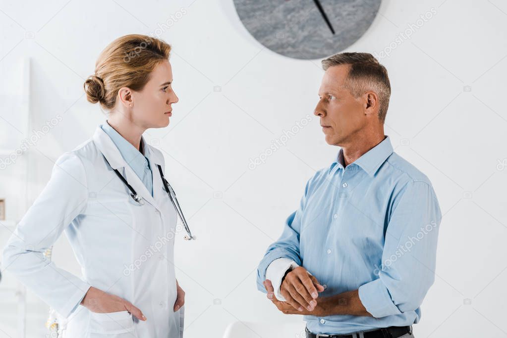 attractive doctor looking at man with broken arm while standing with hands in pockets  in clinic 