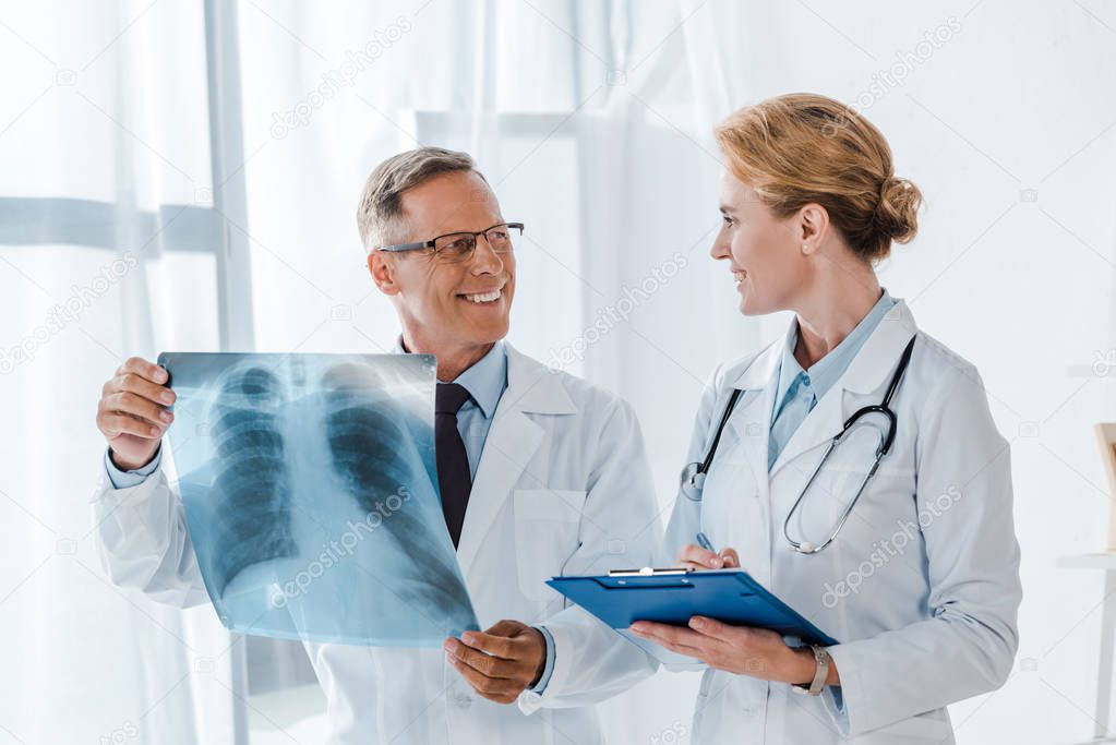 happy doctor holding clipboard and looking at cheerful coworker with x-ray 