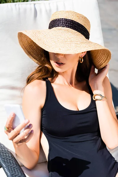 女孩在泳衣和草帽躺在日光浴床上 并使用智能手机在度假村 — 图库照片