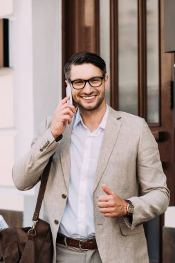 akıllı telefon üzerinde konuşan ve sokakta başparmak gösteren deri çanta ile gülümseyen işadamı ön görünümü