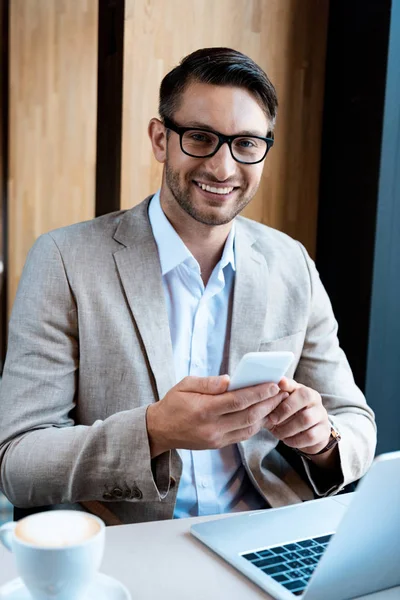 Uśmiechnięty Biznesmen Okularach Posiadających Smartfon Siedząc Przy Stole Laptopem Kawiarni — Zdjęcie stockowe