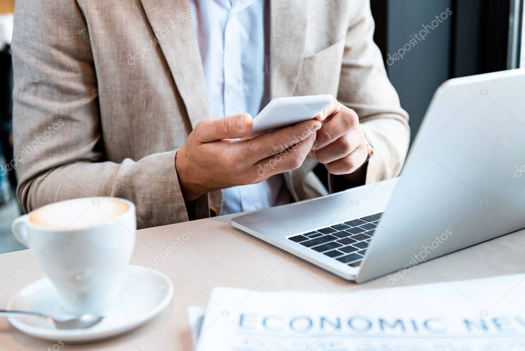partial view of businessman holding smartphone while sitting at table with laptop in cafe