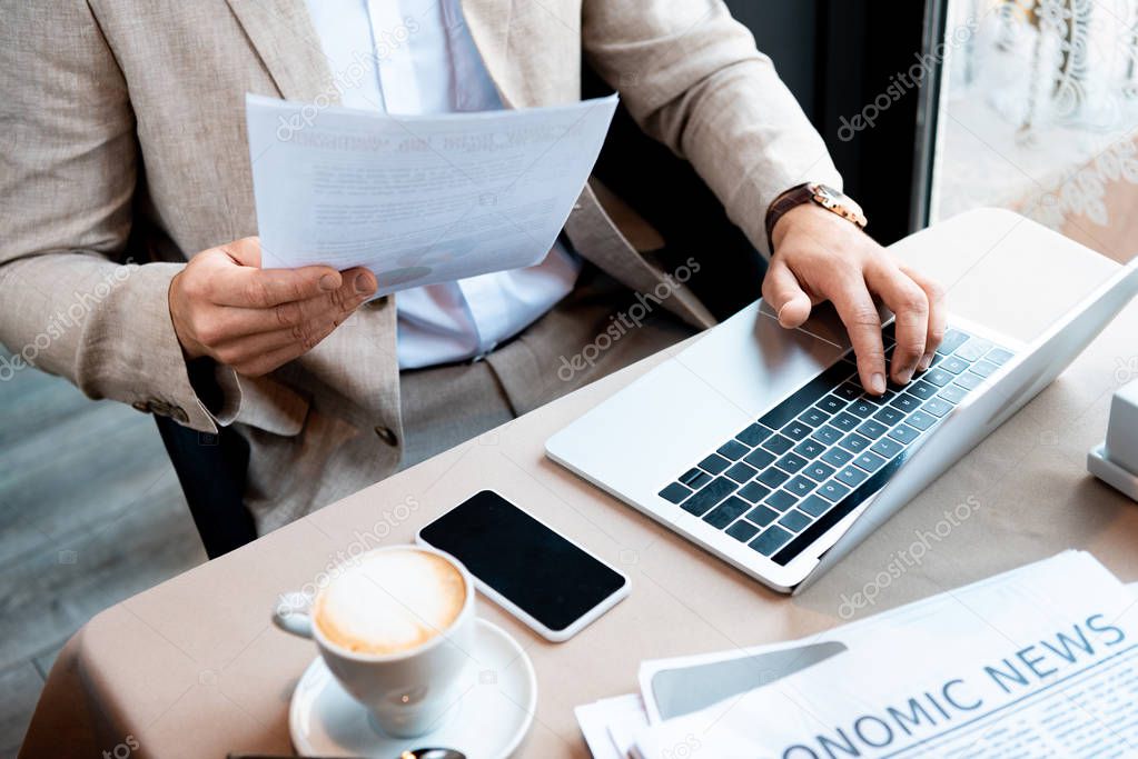 cropped view of businessman holding document and typing on laptop keyboard in cafe