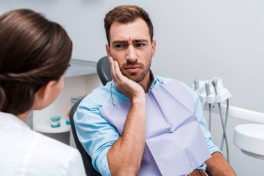 selective focus of upset patient touching face while having toothache near dentist  clipart