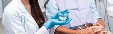 panoramic shot of dentist in latex gloves holding teeth model near patient with clenched hands  clipart
