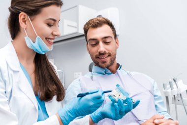 selective focus of attractive dentist in latex gloves holding pen near teeth model and happy patient  clipart