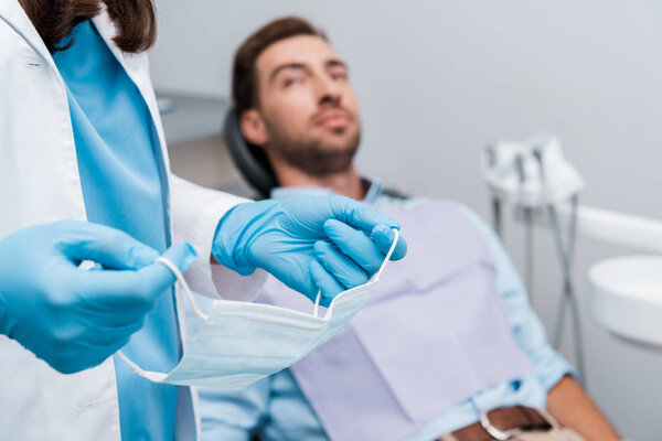 обрезанный вид стоматолога, держащего медицинскую маску возле пациента в стоматологической клинике
 