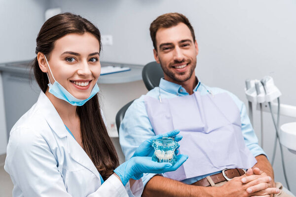 selective focus of attractive dentist in latex gloves holding teeth model near patient with clenched hands 