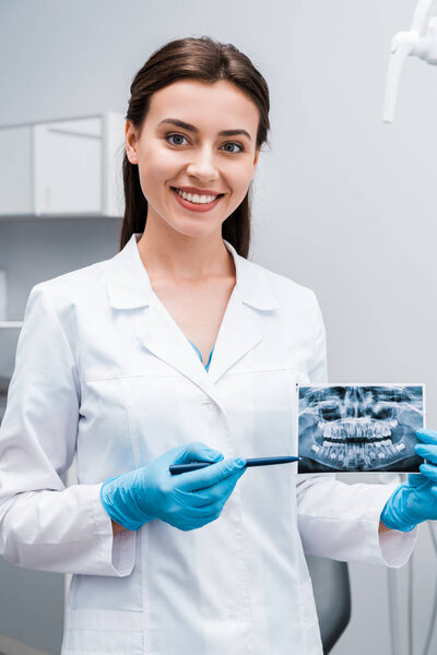 cheerful dentist holding pen near x-ray and smiling in clinic 