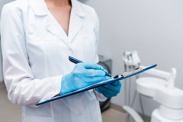 cropped view of dentist writing while holding clipboard in clinic 