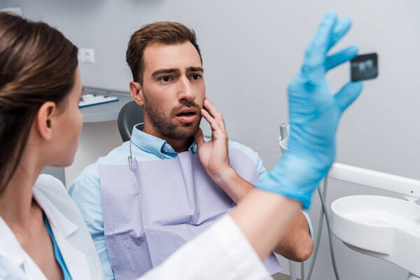 selective focus of scared man touching face and looking at x-ray near dentist 