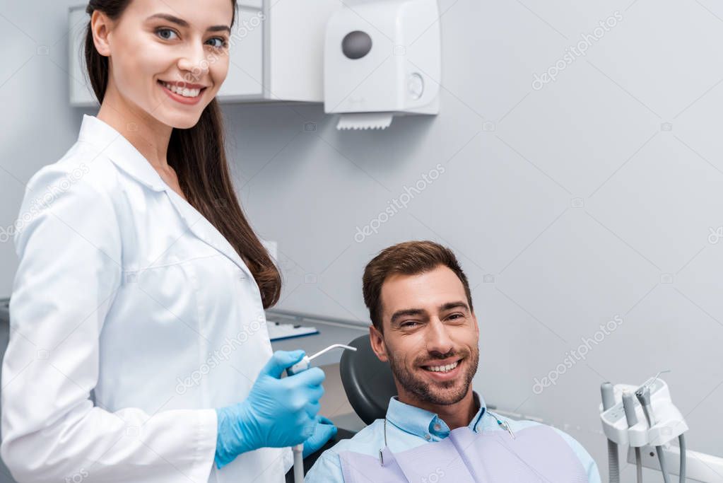 attractive and happy dentist holding dental equipment near happy man 